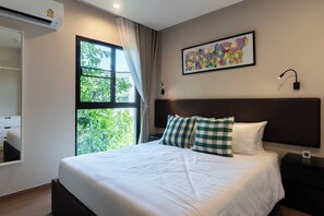 A hotel room with a bed, a window with curtains, a wall-mounted artwork, and a bedside table with a lamp. - Suite Lumaire 1br | Pool view | the title v rawai (Rawai)