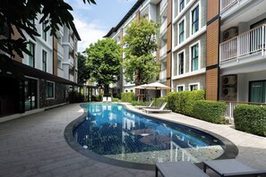 A modern residential building with a swimming pool, lounge chairs, and a covered seating area.