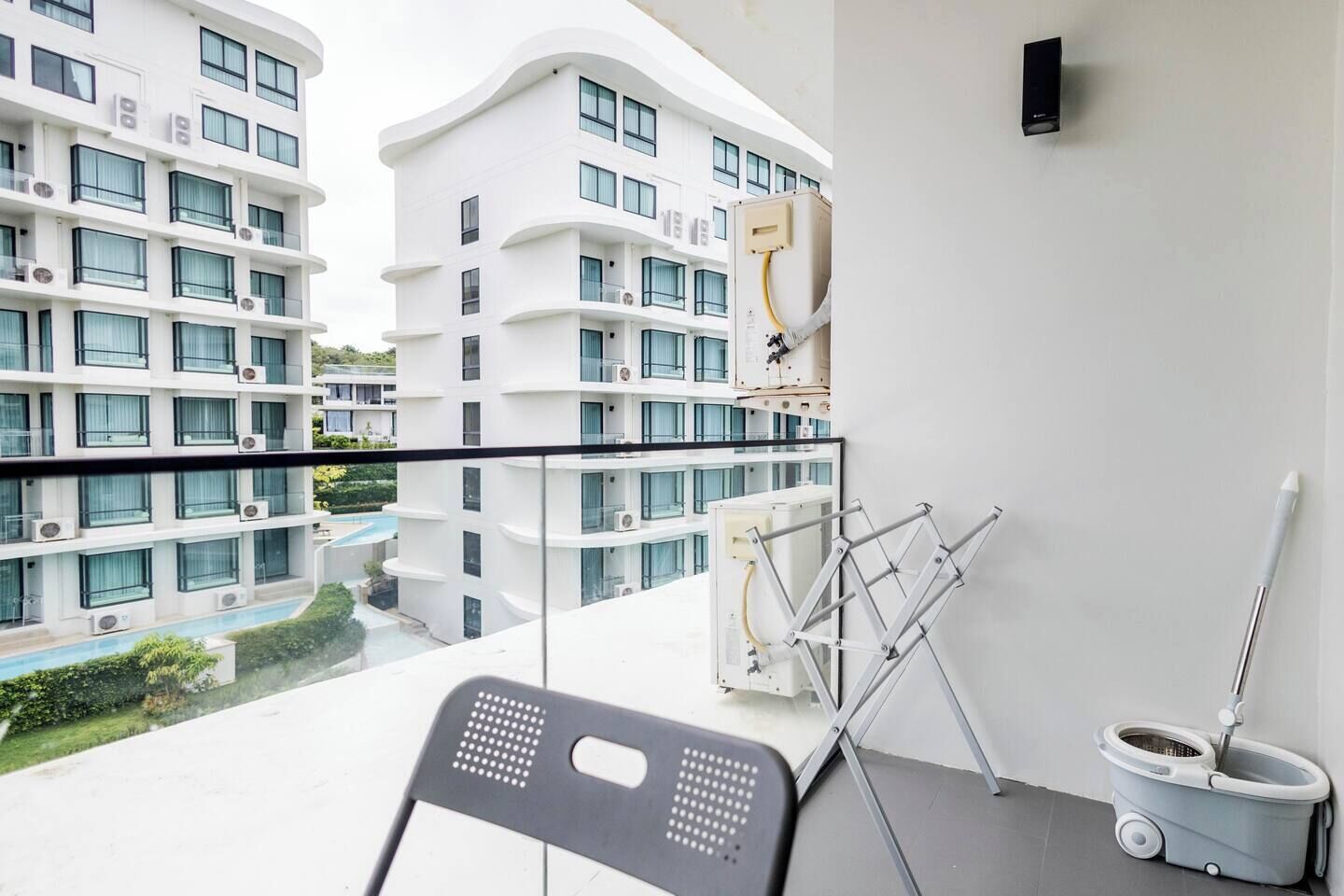 A balcony with a view of a pool and adjacent buildings, a chair, a mop, and a wall-mounted speaker.