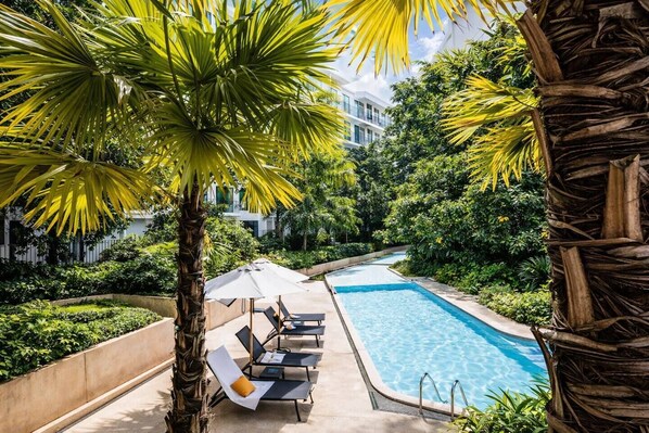 Una piscina con sillones de descanso y sombrillas, rodeada de exuberante vegetación y un edificio moderno al fondo.