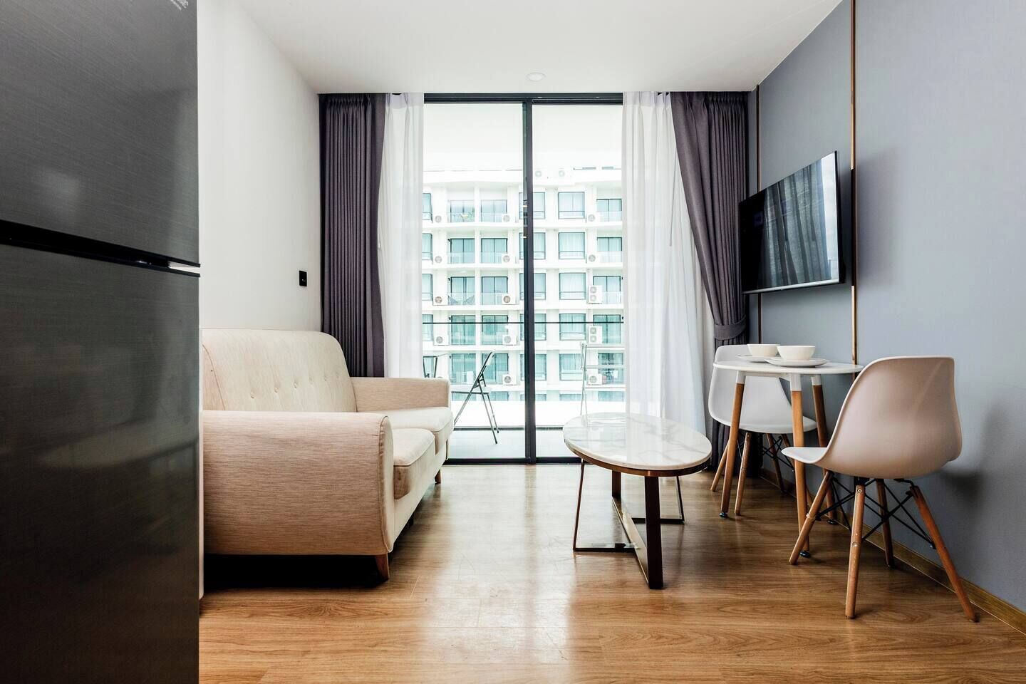 Ein modernes Wohnzimmer mit einer Couch, einem Esstisch mit Stühlen, einem flachbildfernseher an der Wand und einem großen Fenster mit Vorhängen.