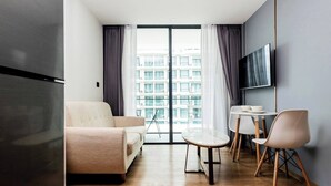 Een moderne woonkamer met een bank, een eettafel met stoelen, een flatscreen-televisie aan de muur en een groot raam met gordijnen.