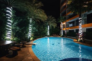 A nighttime swimming pool with surrounding palm trees and a building with orange and white exterior.