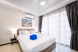 A hotel room with a bed, a nightstand, a lamp, an air conditioner, a painting, and a window with curtains. - Nova Condo 1br | Pool view | nbc nai harn (Rawai)