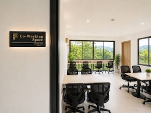 A co-working space with multiple tables and chairs, large windows with a view of greenery, and a sign indicating the space name.