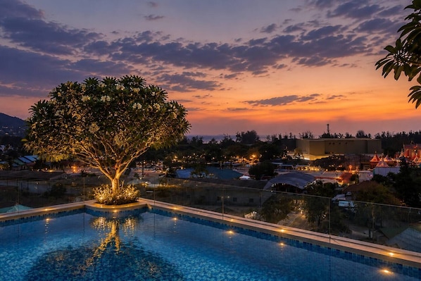 A swimming pool with a tree and lights, surrounded by buildings and a sunset.