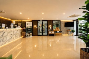 A hotel reception area with a marble counter, a reception desk, a large poster, and a reception desk with a computer and a plant.