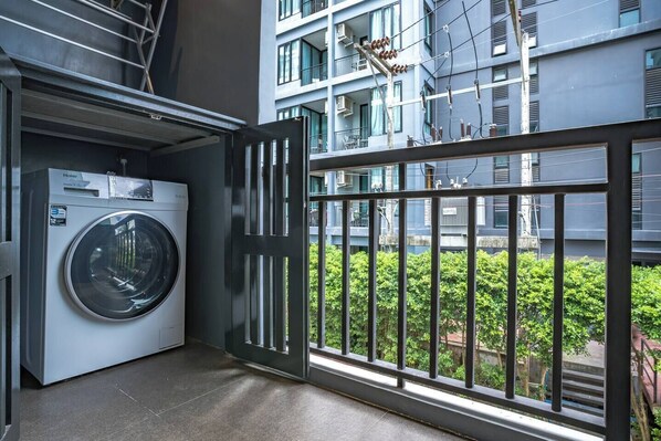 A laundry room with a front-loading washer and dryer, a metal railing, and a view of a residential building with balconies.