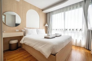 Een moderne hotelkamer met een houten vloer, een bed met witte beddengoed, een ronde spiegel, een nachtkastje en een raam met gordijnen.