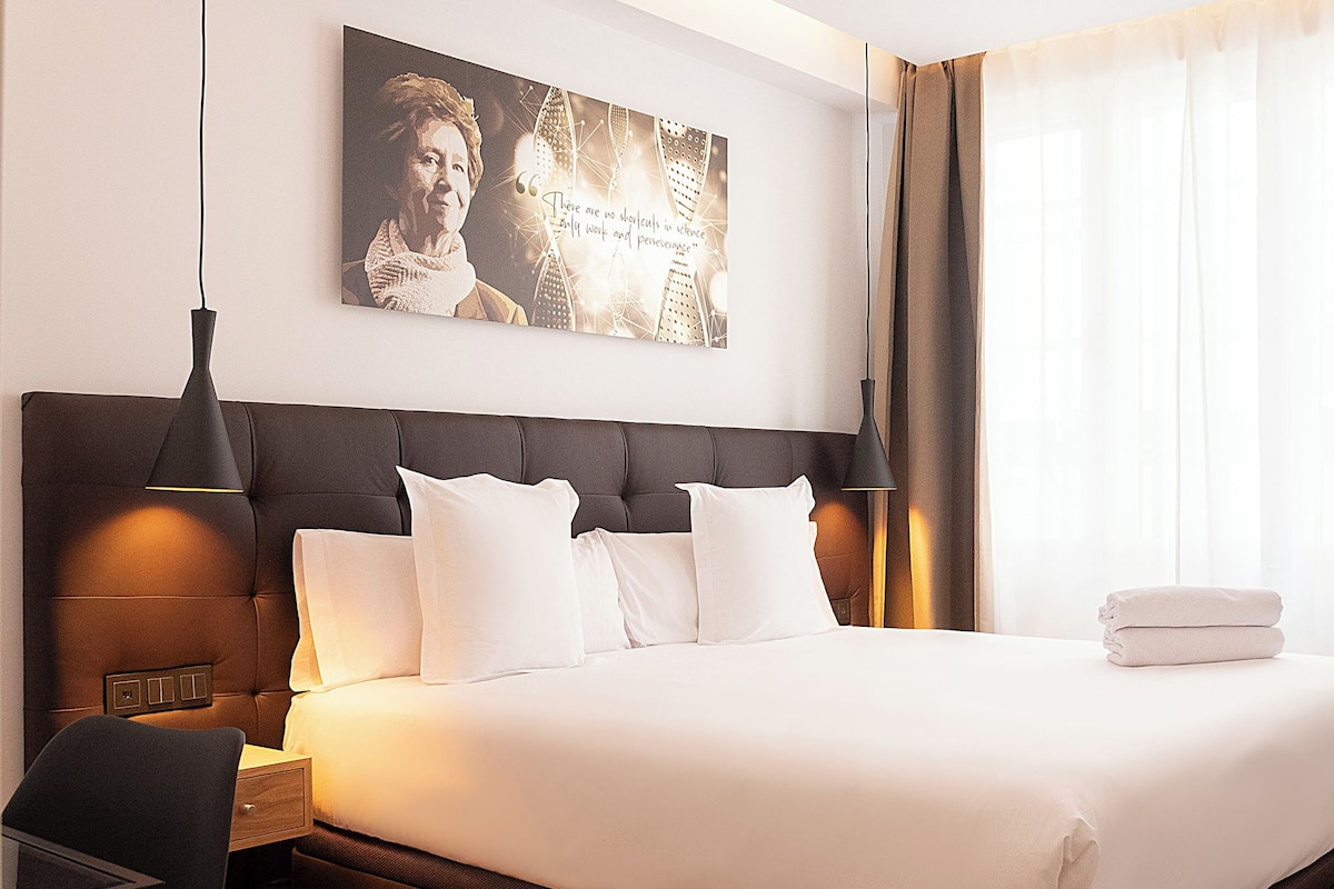 Une chambre d’hôtel avec un grand lit, une tête de lit, une table de chevet, une lampe, un tableau au mur et une fenêtre avec des rideaux.