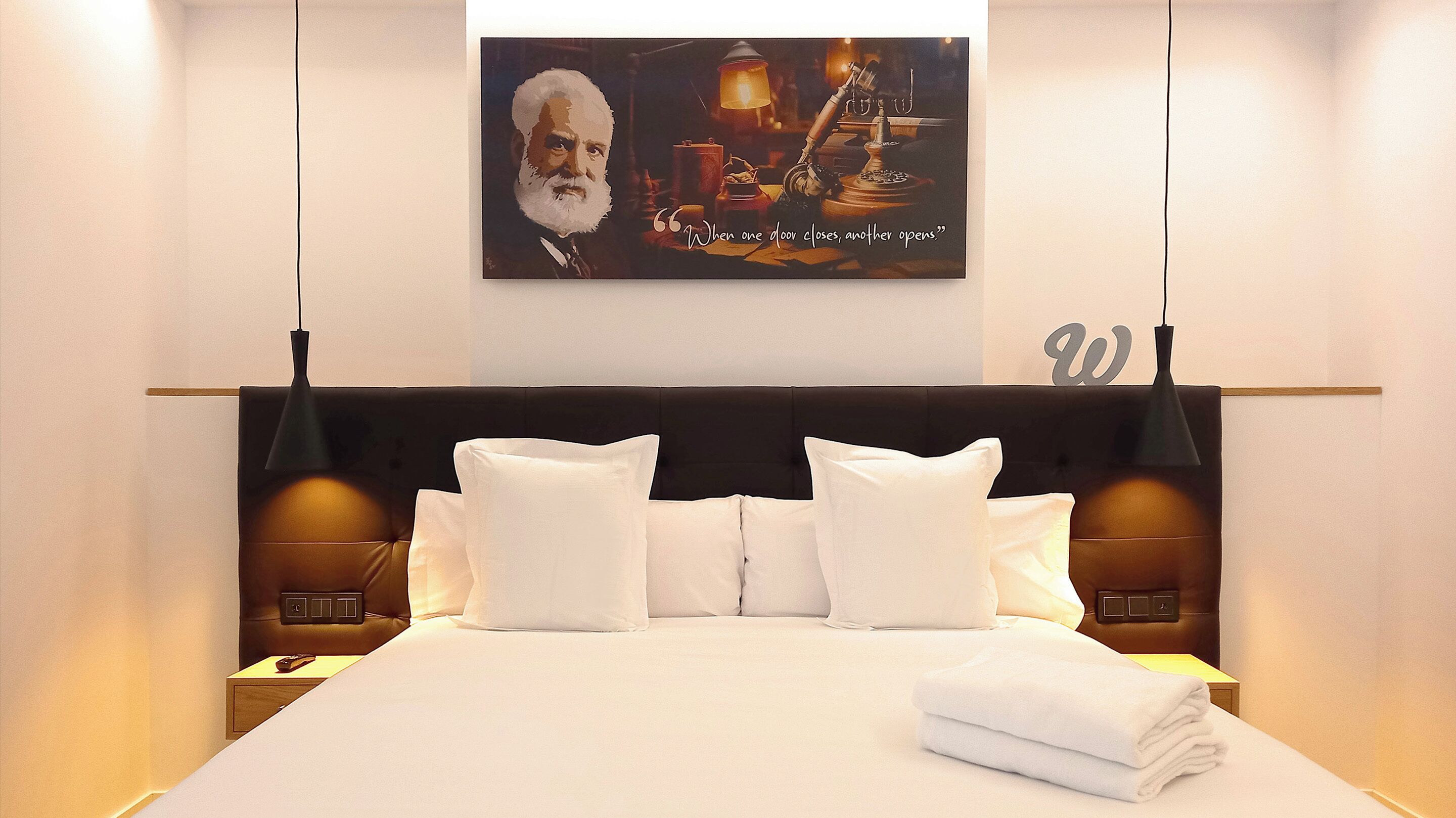 Une chambre d’hôtel avec un grand lit, du linge de lit blanc, deux lampes de chevet, une citation encadrée au mur et un élément décoratif en forme de lettre « W ».