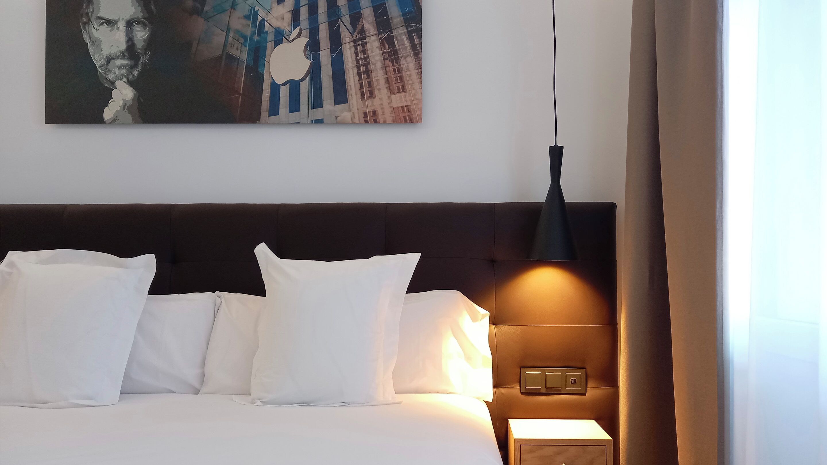 Habitación de hotel con cama, almohadas blancas, cabecera oscura, lámpara de pared y un cuadro sobre la cama.