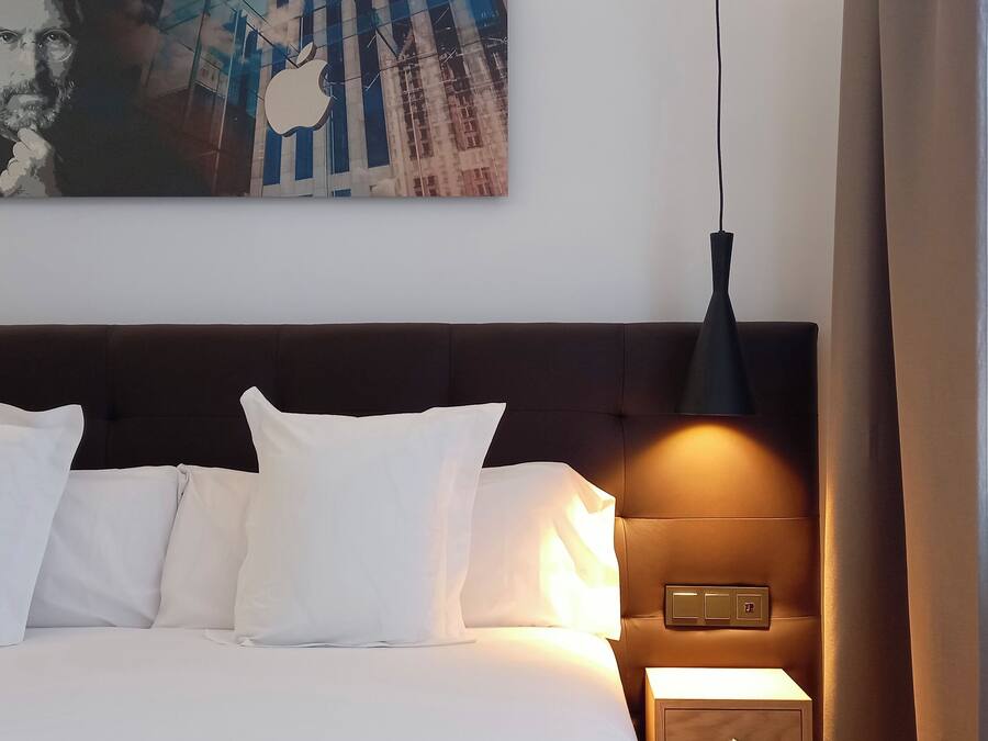 Habitación de hotel con cama, almohadas blancas, cabecera oscura, lámpara de pared y un cuadro sobre la cama.