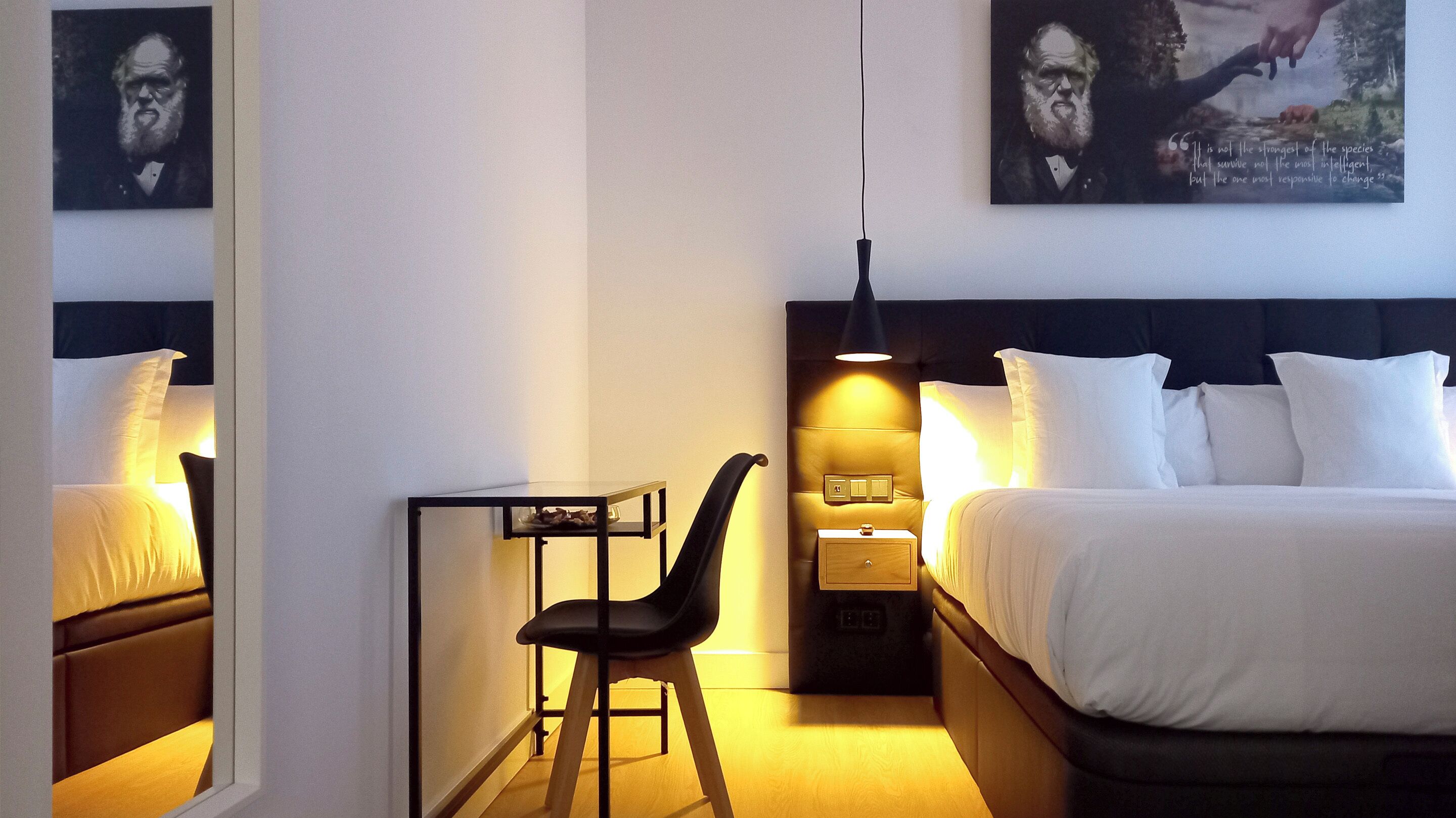 A hotel room with a bed, a desk, a chair, a nightstand, and a wall-mounted lamp.