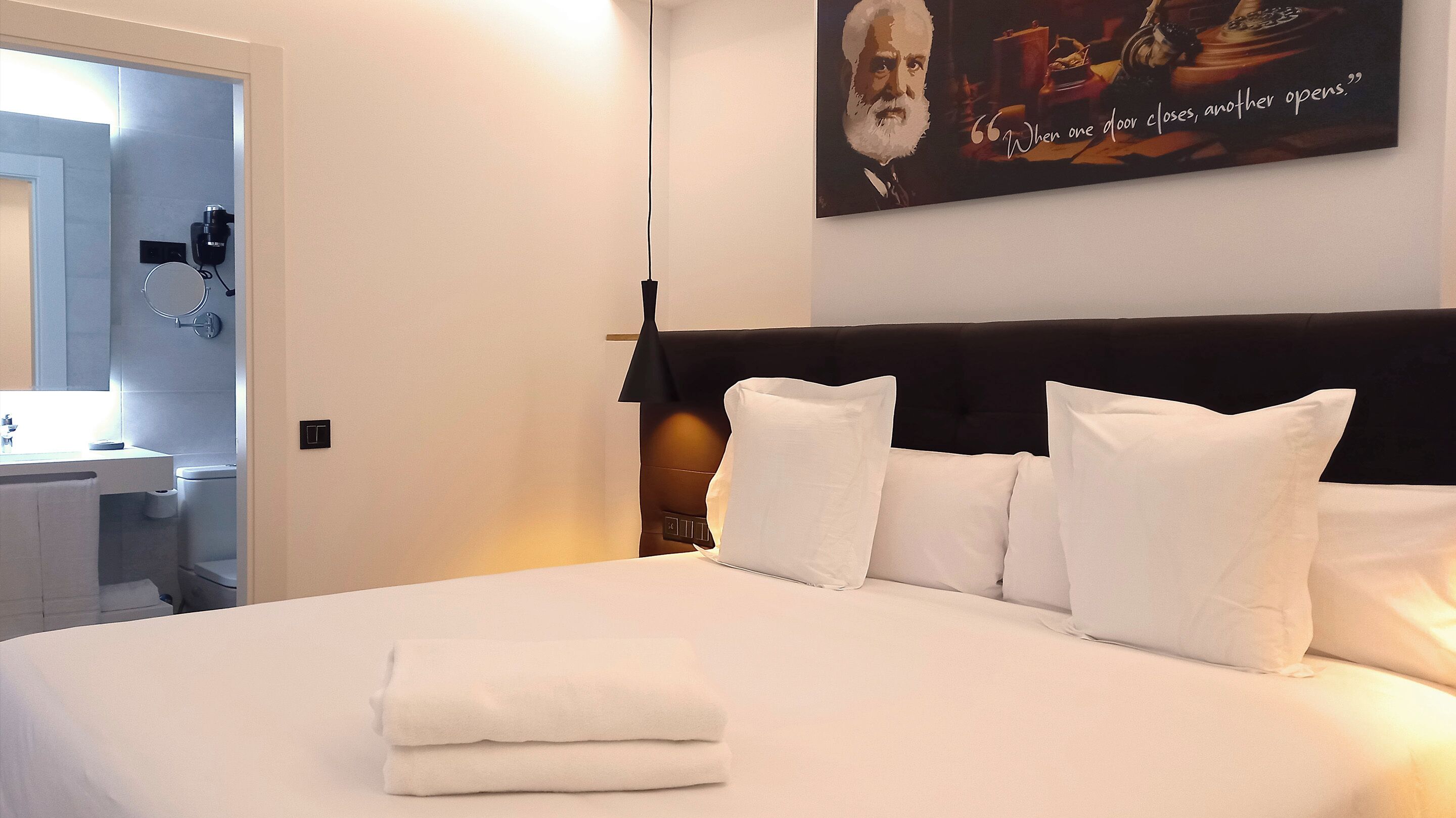 A hotel room with a neatly made bed, a bedside table, a wall-mounted lamp, and a poster on the wall.
