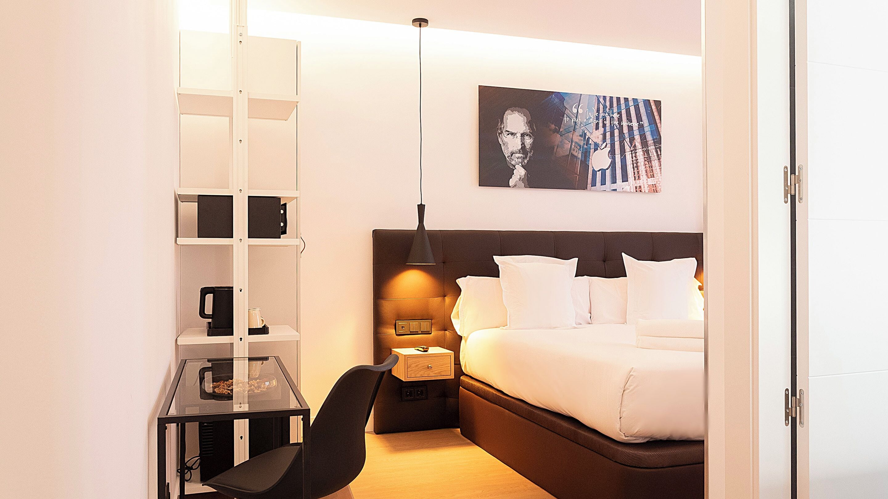 Habitación de hotel con cama, escritorio y silla, estantería, mesita de noche con lámpara y una obra de arte en la pared.