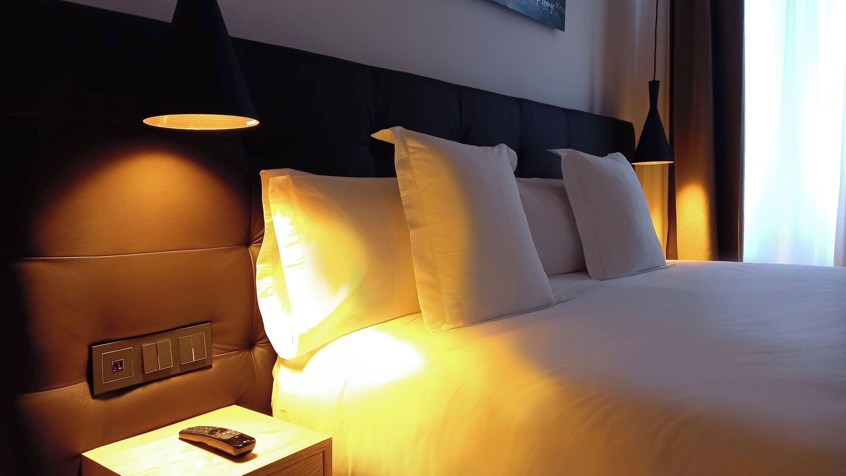 A hotel room with a large bed, multiple pillows, a nightstand with a remote control, and wall-mounted lamps.