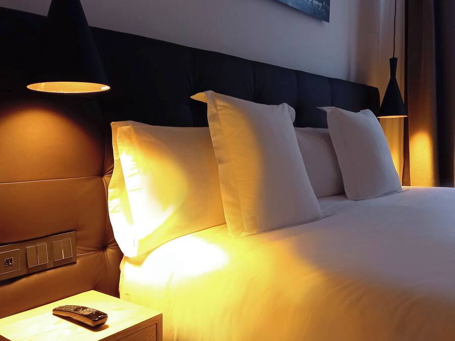 A hotel room with a large bed, multiple pillows, a nightstand with a remote control, and wall-mounted lamps.