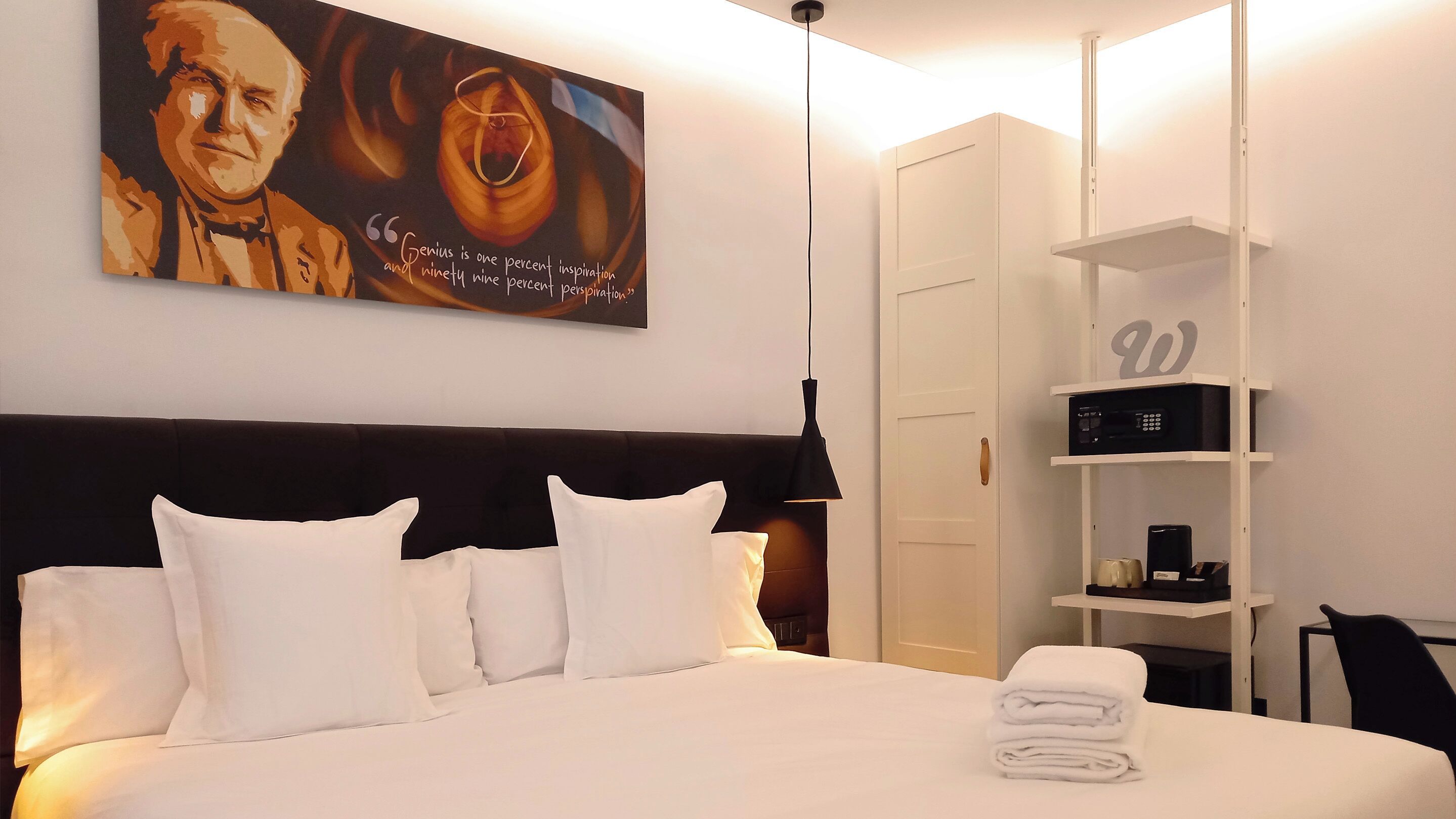 Habitación de hotel con una cama grande, almohadas blancas, cabecero, un cuadro en la pared, armario, estante con objetos y escritorio con silla.