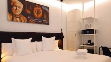 Habitación de hotel con una cama grande, almohadas blancas, cabecero, un cuadro en la pared, armario, estante con objetos y escritorio con silla.