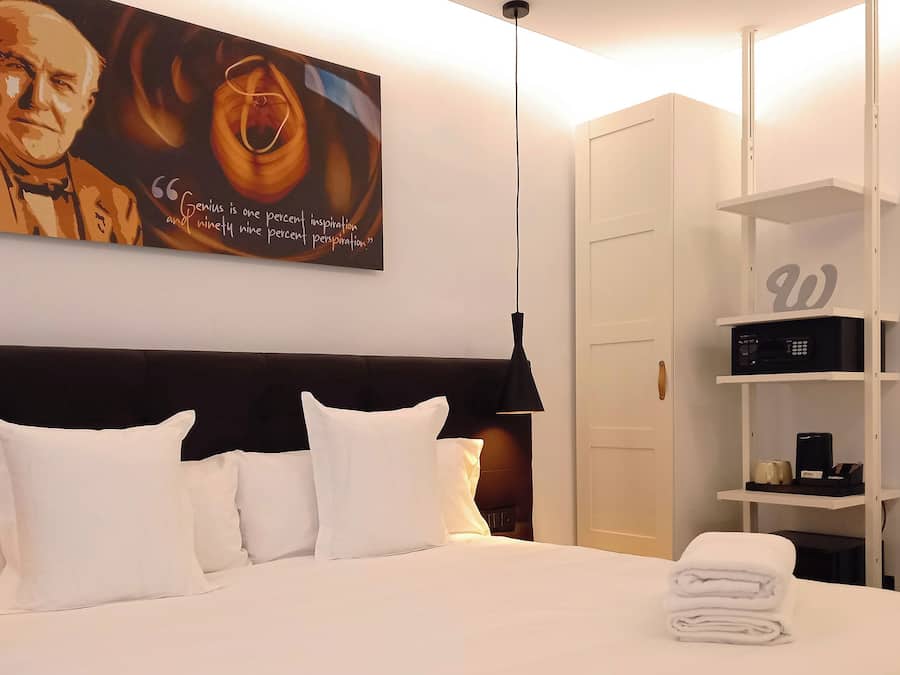 Habitación de hotel con una cama grande, almohadas blancas, cabecero, un cuadro en la pared, armario, estante con objetos y escritorio con silla.