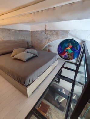 Une chambre à coucher avec un lit, des oreillers, une table en verre et une œuvre d’art aux couleurs vives.