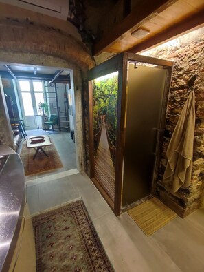 Un couloir avec un mur en pierre rustique, une porte en bois donnant sur un paysage verdoyant, une petite table avec un bol, et un tapis à motifs.