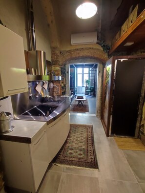 Une cuisine équipée d’un évier en acier inoxydable, d’une cuisinière et d’un porte-ustensiles suspendu. Un tapis recouvre le sol et une porte donne accès à une autre pièce.