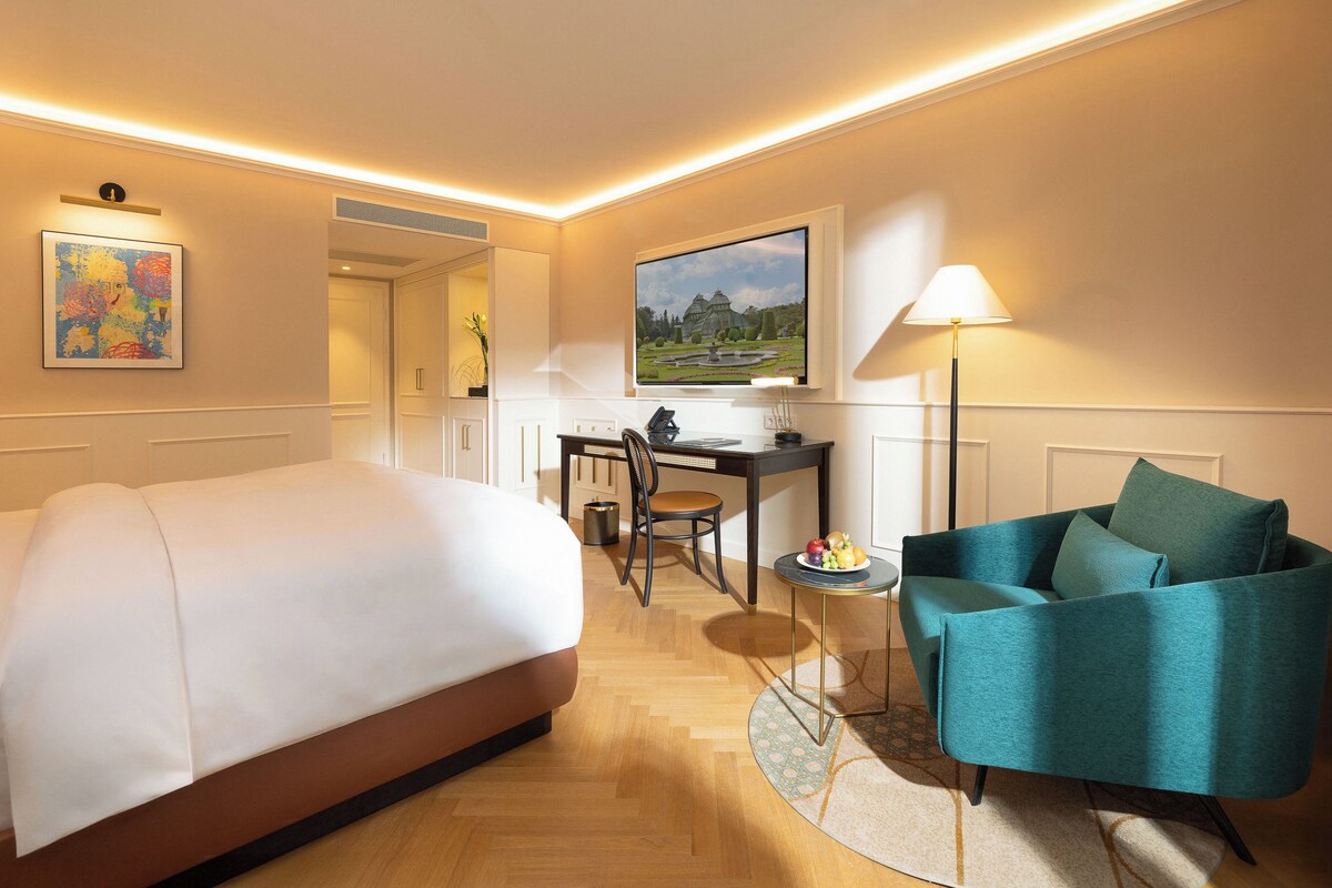 A hotel room with a bed, a desk, a teal armchair, a TV, and a lamp.
