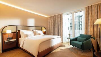 A hotel room with a large bed, a nightstand with a lamp, a green armchair, and a view of the city through large windows.