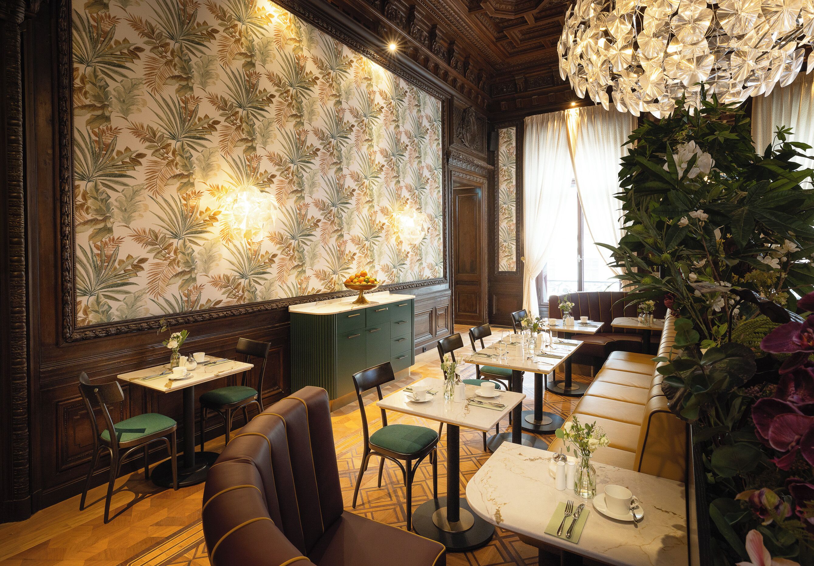 A restaurant with wooden paneling, green upholstered chairs, and a patterned wallpaper. Tables are set with white tablecloths and floral centerpieces.