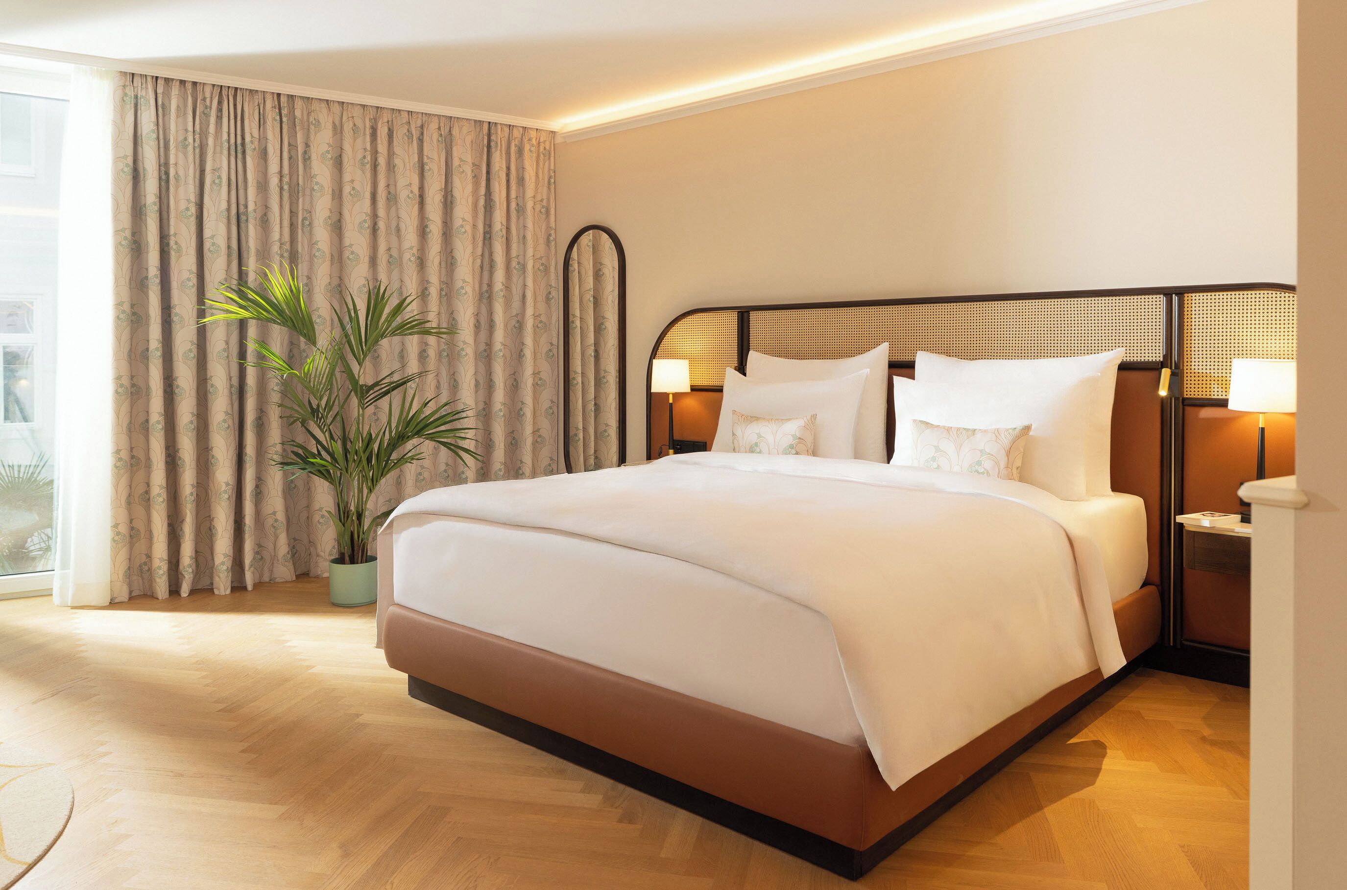 Ein Hotelzimmer mit einem großen Bett, einer Zimmerpflanze, einem Fenster mit Vorhängen und Nachttischlampen.