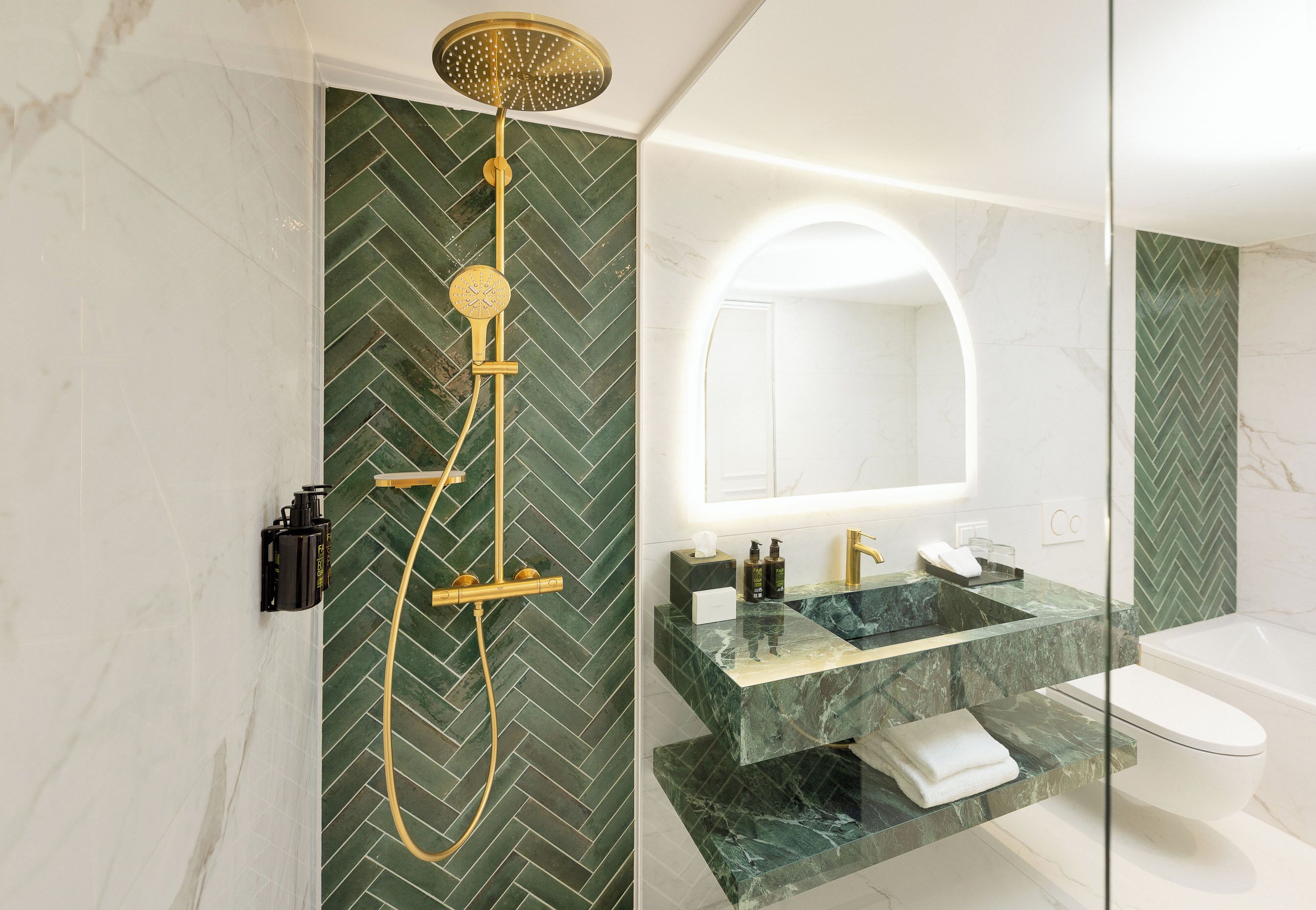 Uma casa de banho moderna com uma parede verde em padrão herringbone, um chuveiro dourado, uma pia de mármore e uma sanita branca.