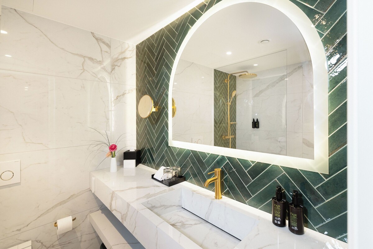 A modern bathroom with a marble sink, a large mirror, and a green herringbone tile backsplash.