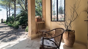 A covered outdoor area with a wicker chair, a large vase with branches, and a window with decorative iron bars.