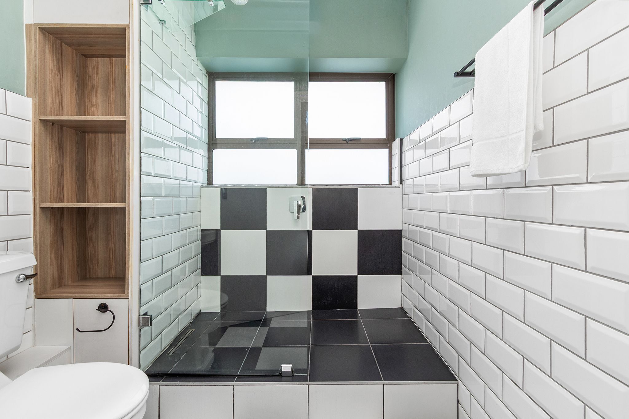 A bathroom with a toilet, a wooden shelf, a black and white tiled shower area, and a window.