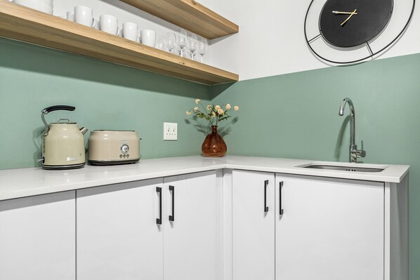 A kitchen with white cabinets, a kettle, a toaster, a vase with flowers, a sink, and a clock on the wall. - 1 Plomarion (Cape Town)