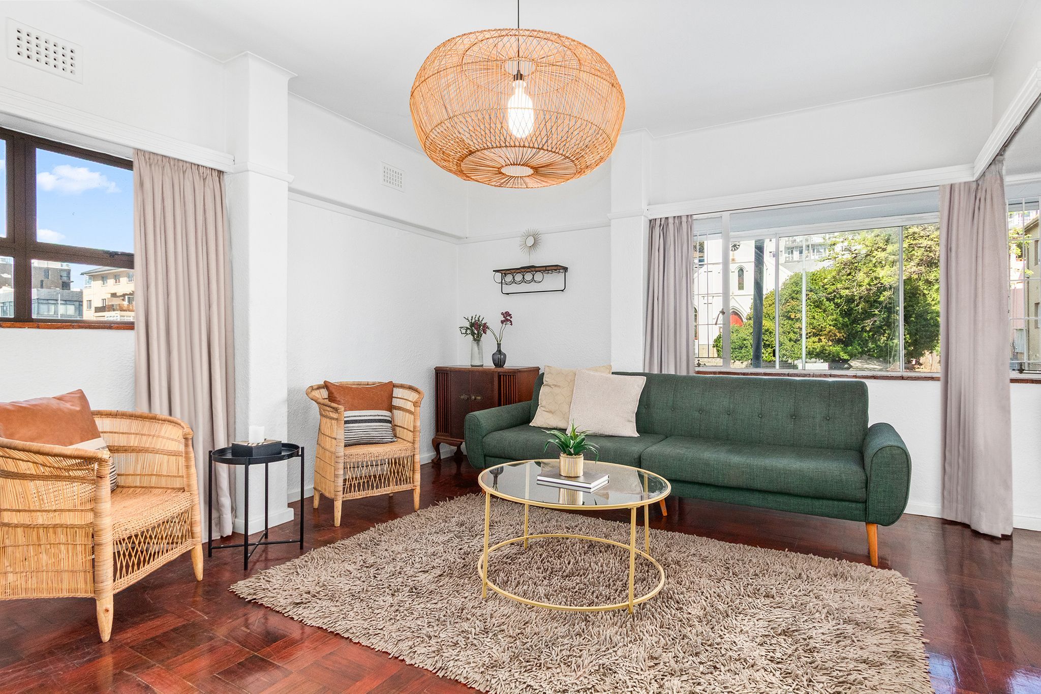 Un salon avec un canapé vert, des fauteuils en osier, une table basse en verre et une grande fenêtre avec des rideaux.