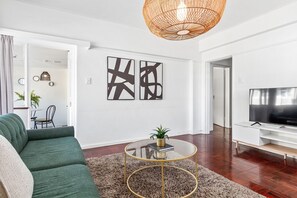 A modern living room with a green sofa, a glass coffee table with a gold base, abstract wall art, a television, and a wicker ceiling light. - 1 Plomarion (Cape Town)