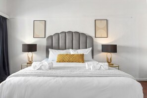 A neatly made bed with a grey upholstered headboard, white bedding, and a single decorative pillow. There are two bedside tables with lamps and a framed artwork above each. - 1 Plomarion (Cape Town)