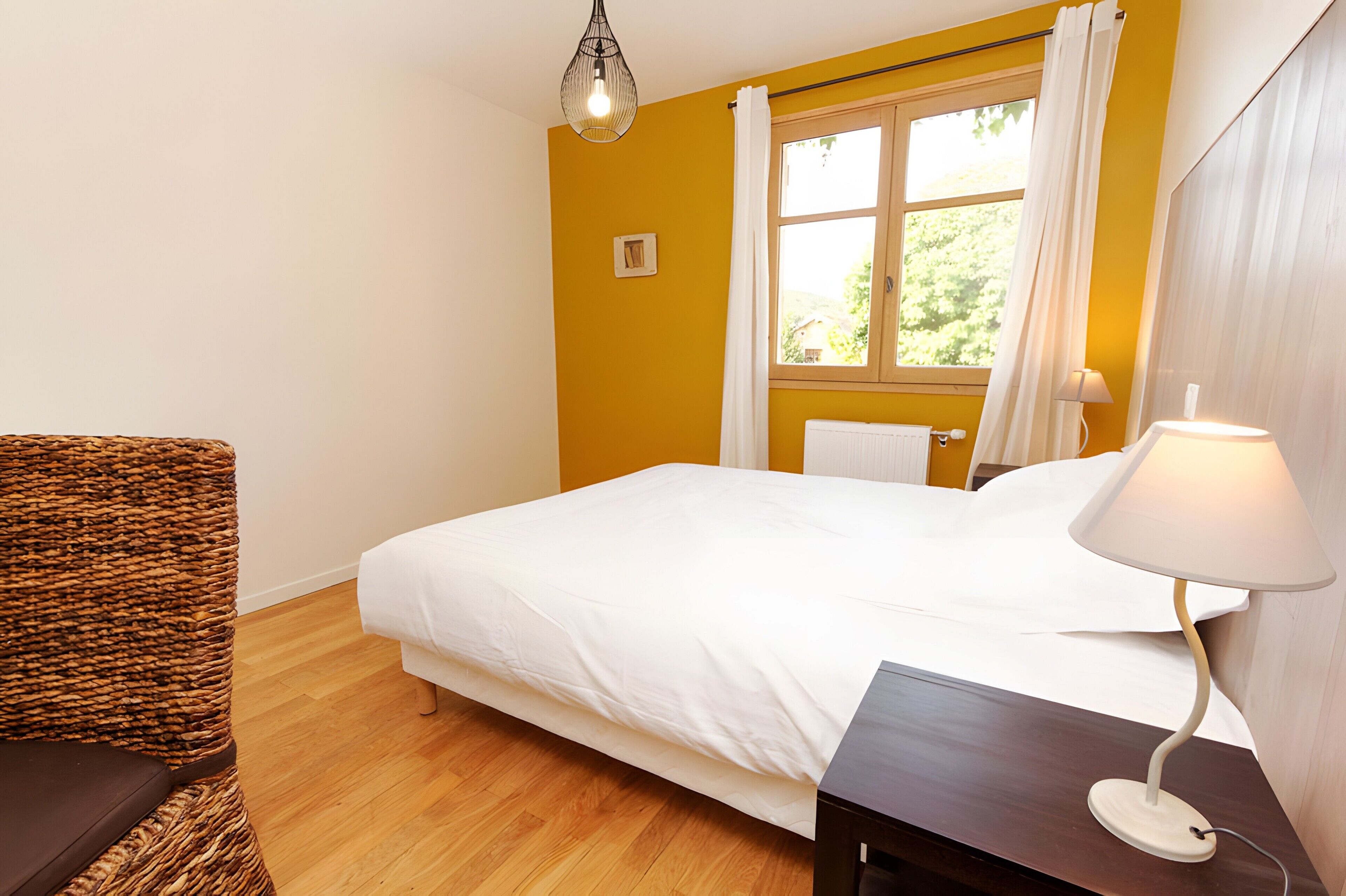 A hotel room with a bed, a chair, a lamp, a window with curtains, and a wooden floor.