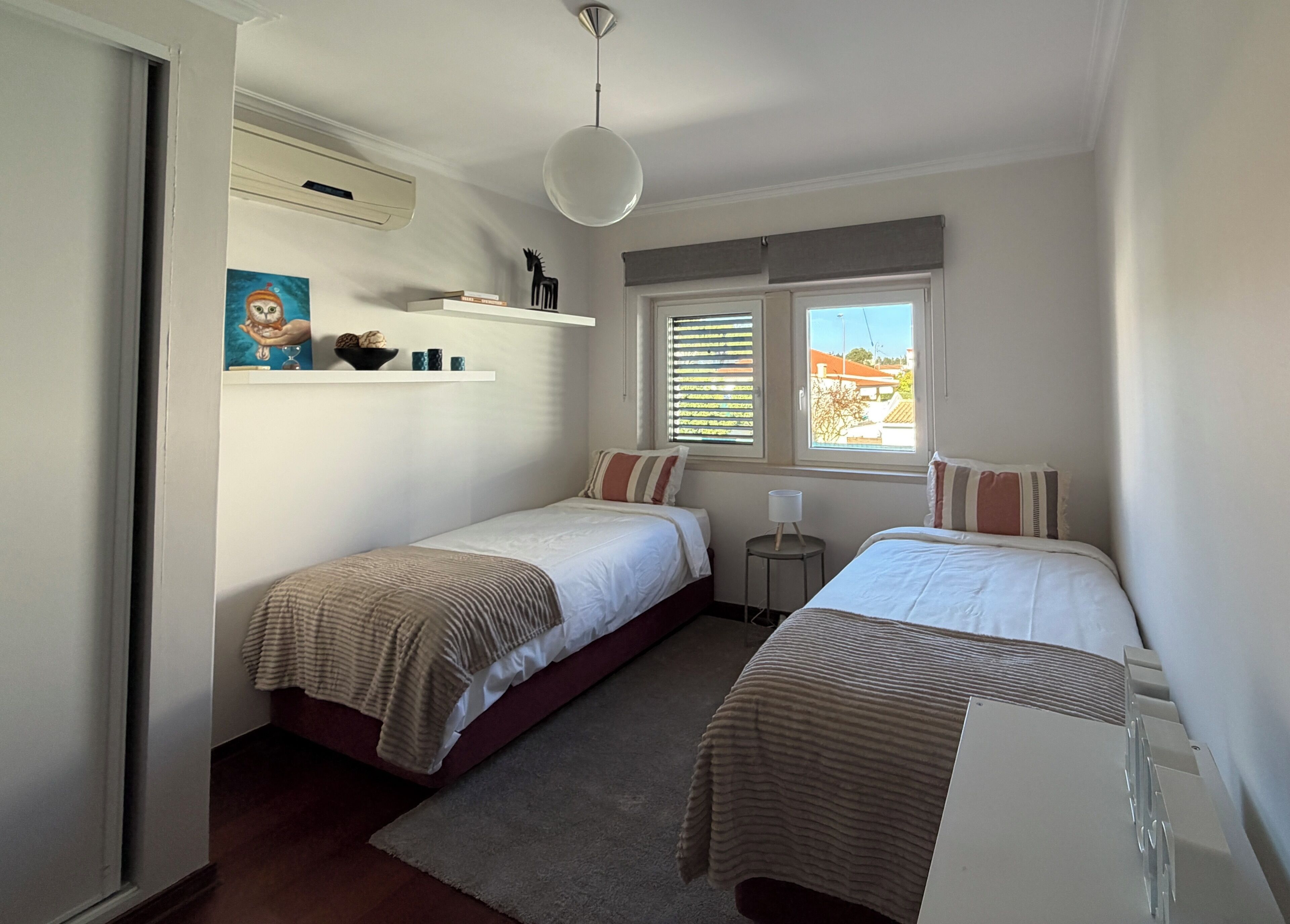 A bedroom with two beds, a small table, a shelf with books and a decorative item, a window with blinds, and a ceiling light.