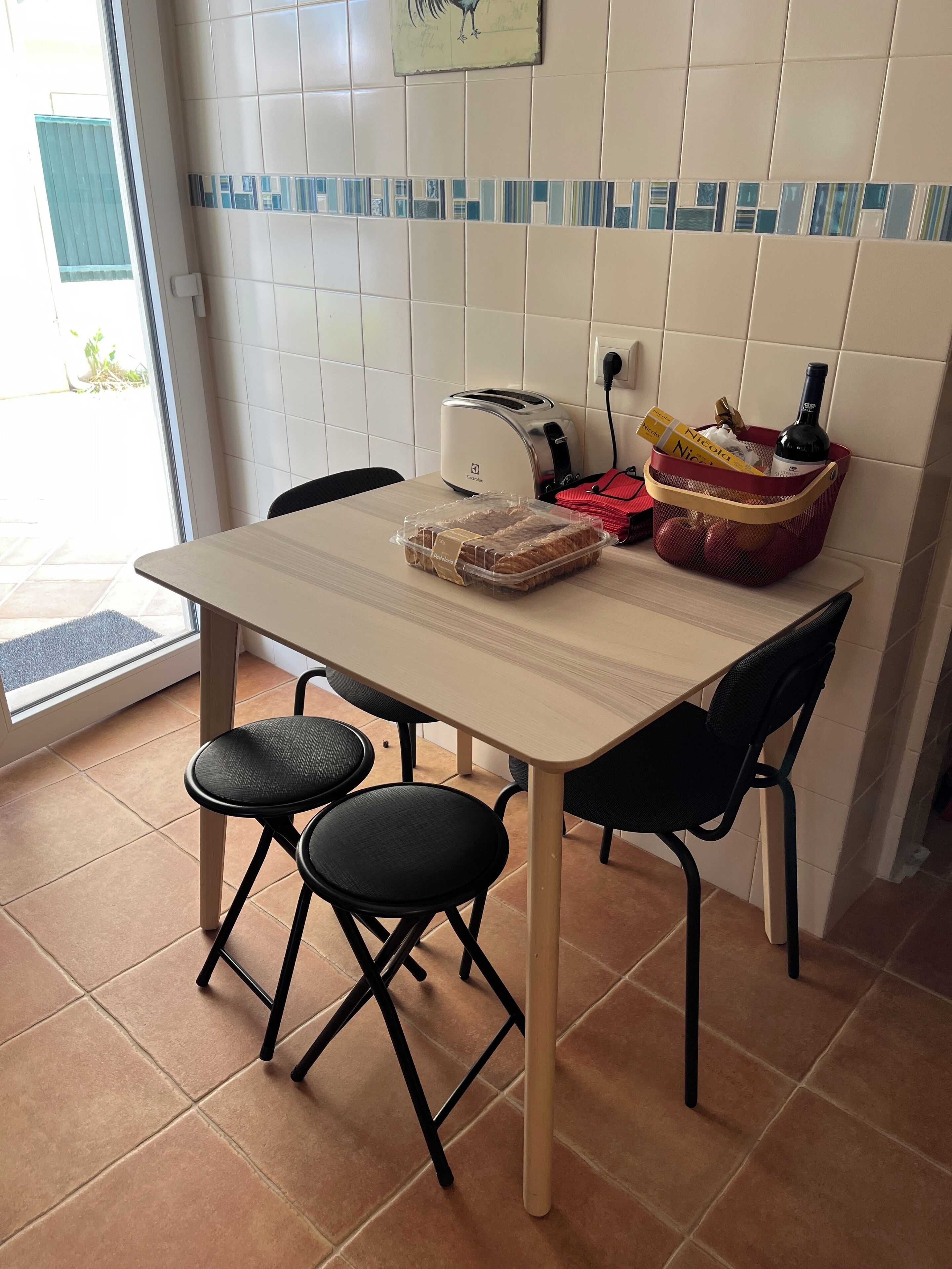 Una cocina con una mesa, sillas, un tostador, una cesta con productos y una ventana.