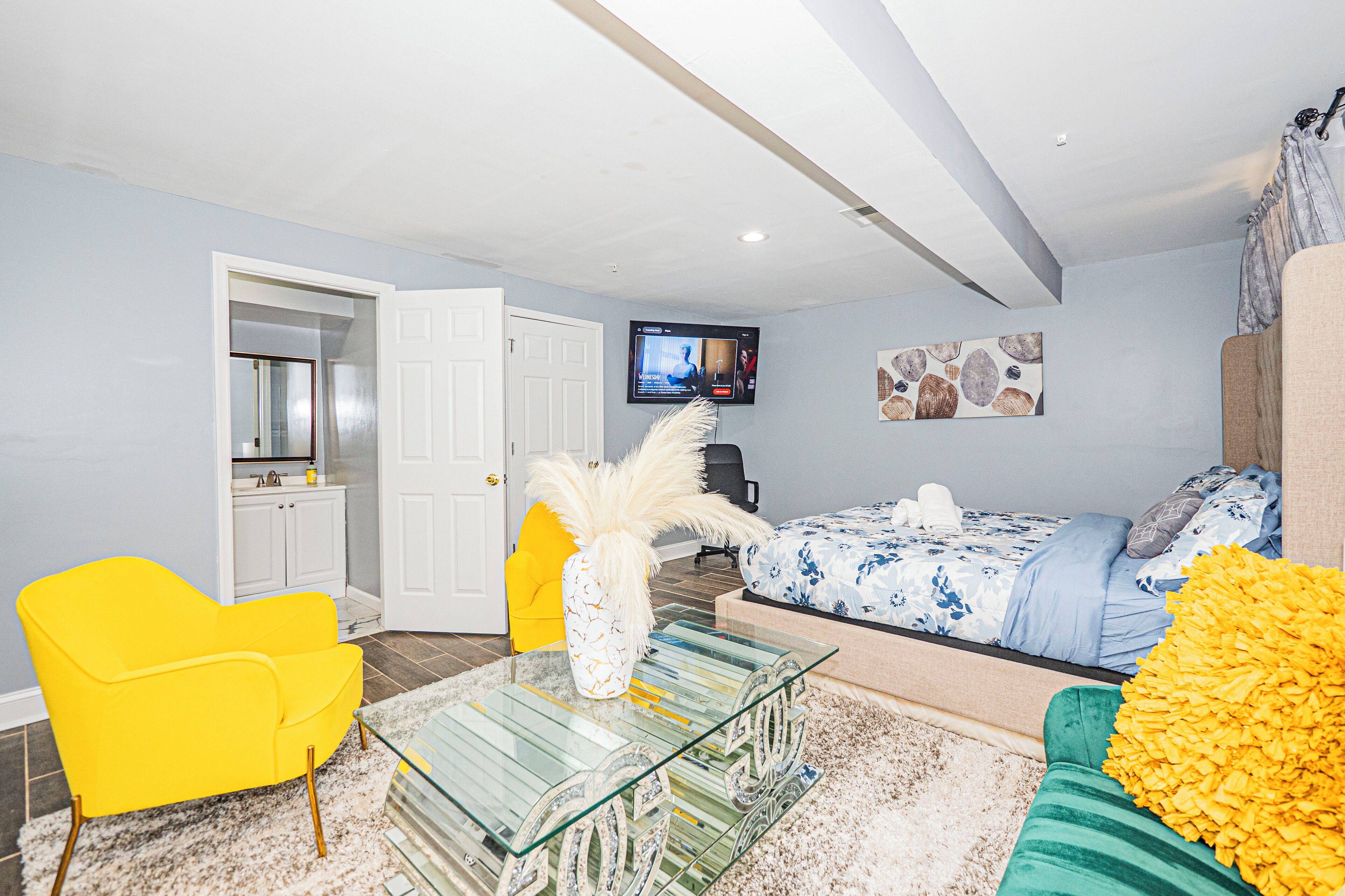A modern living room with a glass coffee table, a yellow armchair, a green sofa, and a bed with blue bedding.