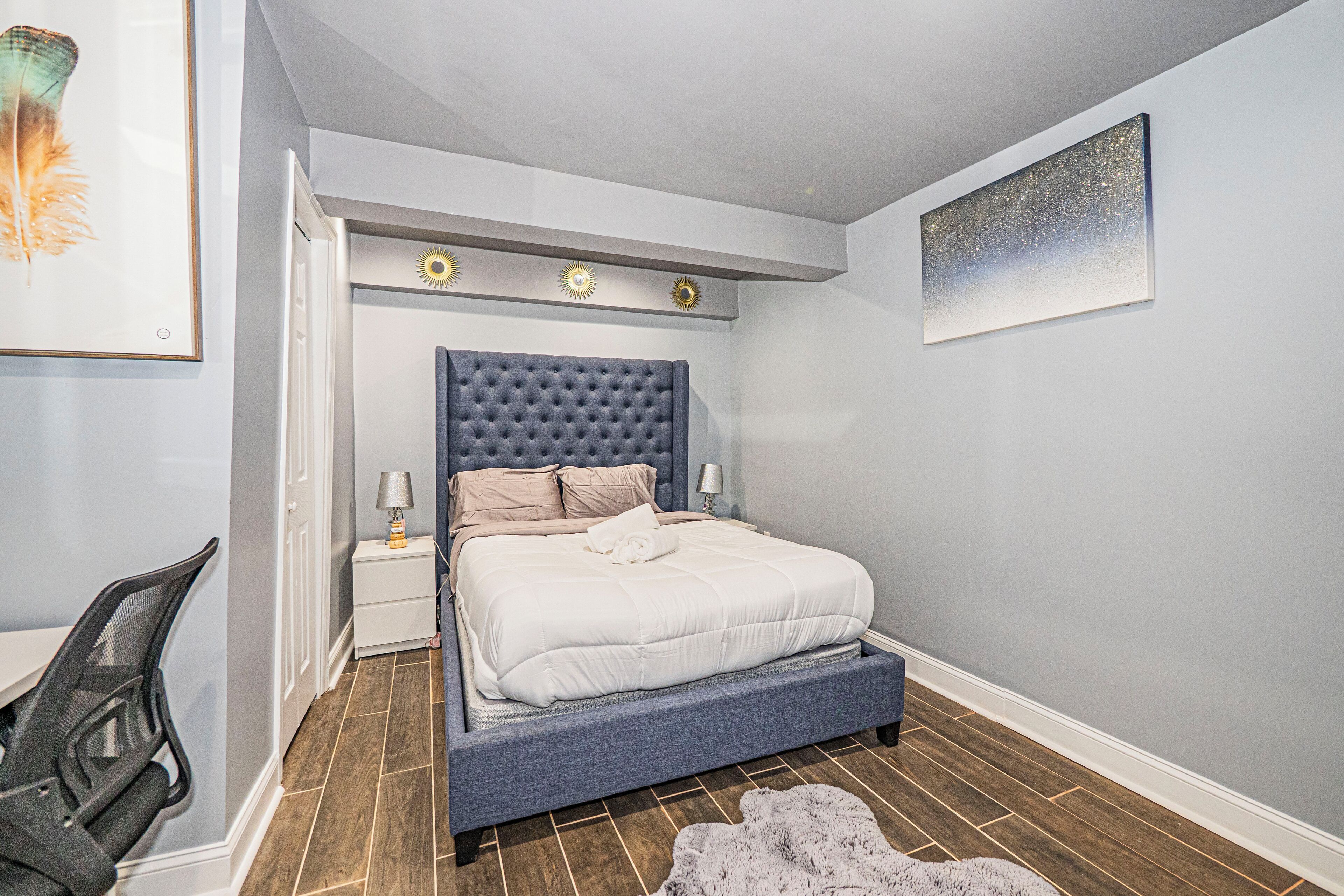 Ein Schlafzimmer mit einer gepolsterten Kopfteile, einem Bett mit weißer Bettwäsche, einem kleinen Nachttisch mit Lampe und einem gerahmten Bild an der Wand.