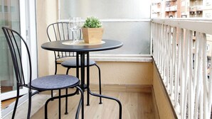 Un balcon avec une table ronde, deux chaises, un pot de fleurs et une vue sur un immeuble d’habitation.