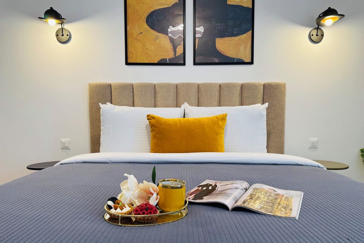 A neatly made bed with a blue blanket, a yellow pillow, and a tray with snacks and a drink. There are two wall-mounted lamps and two framed artworks above the bed.