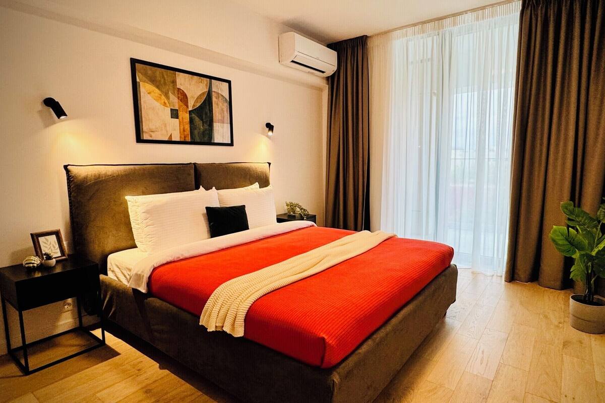 A hotel room with a large bed, a nightstand, a potted plant, and a painting on the wall.