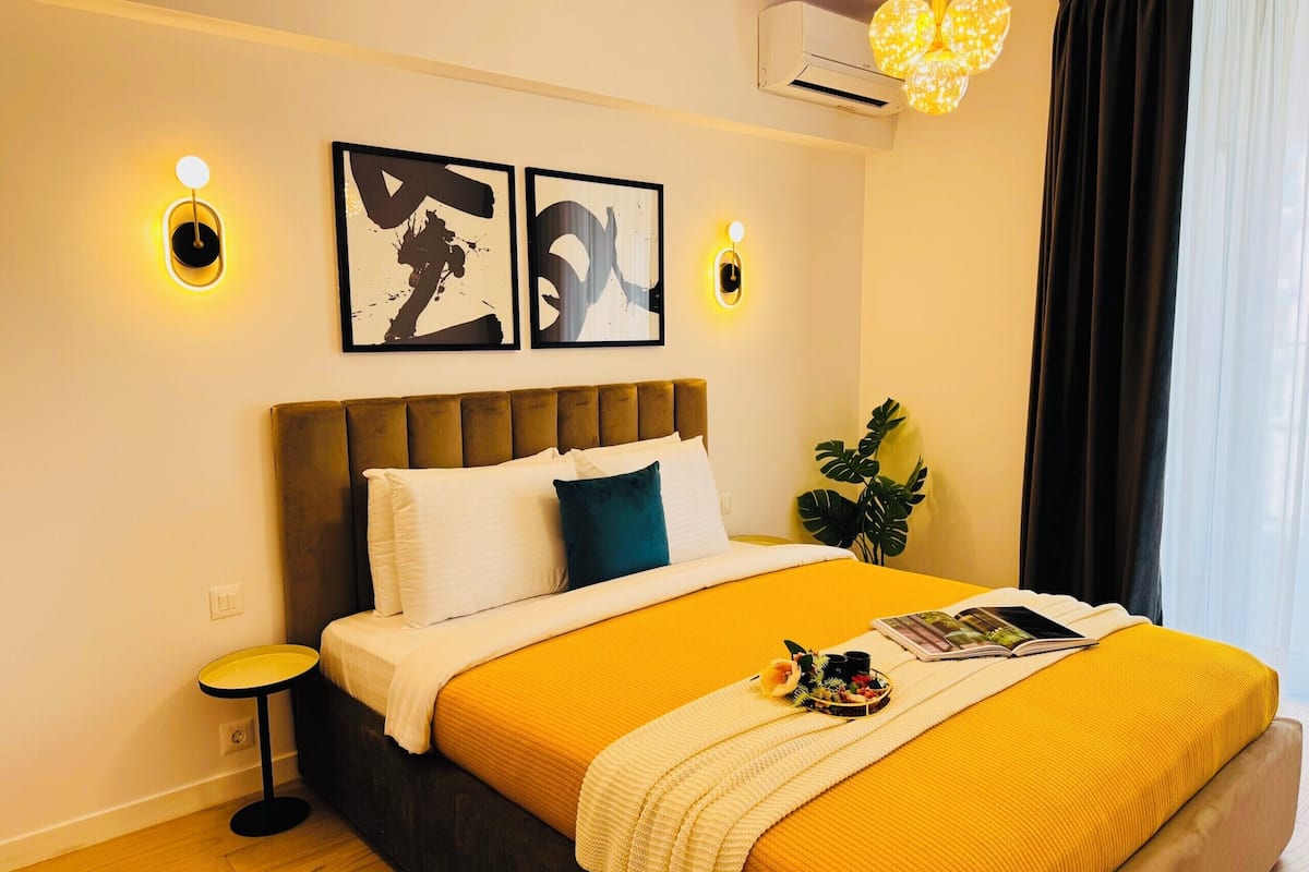 A hotel room with a bed, a bedside table, a plant, and artwork on the wall.