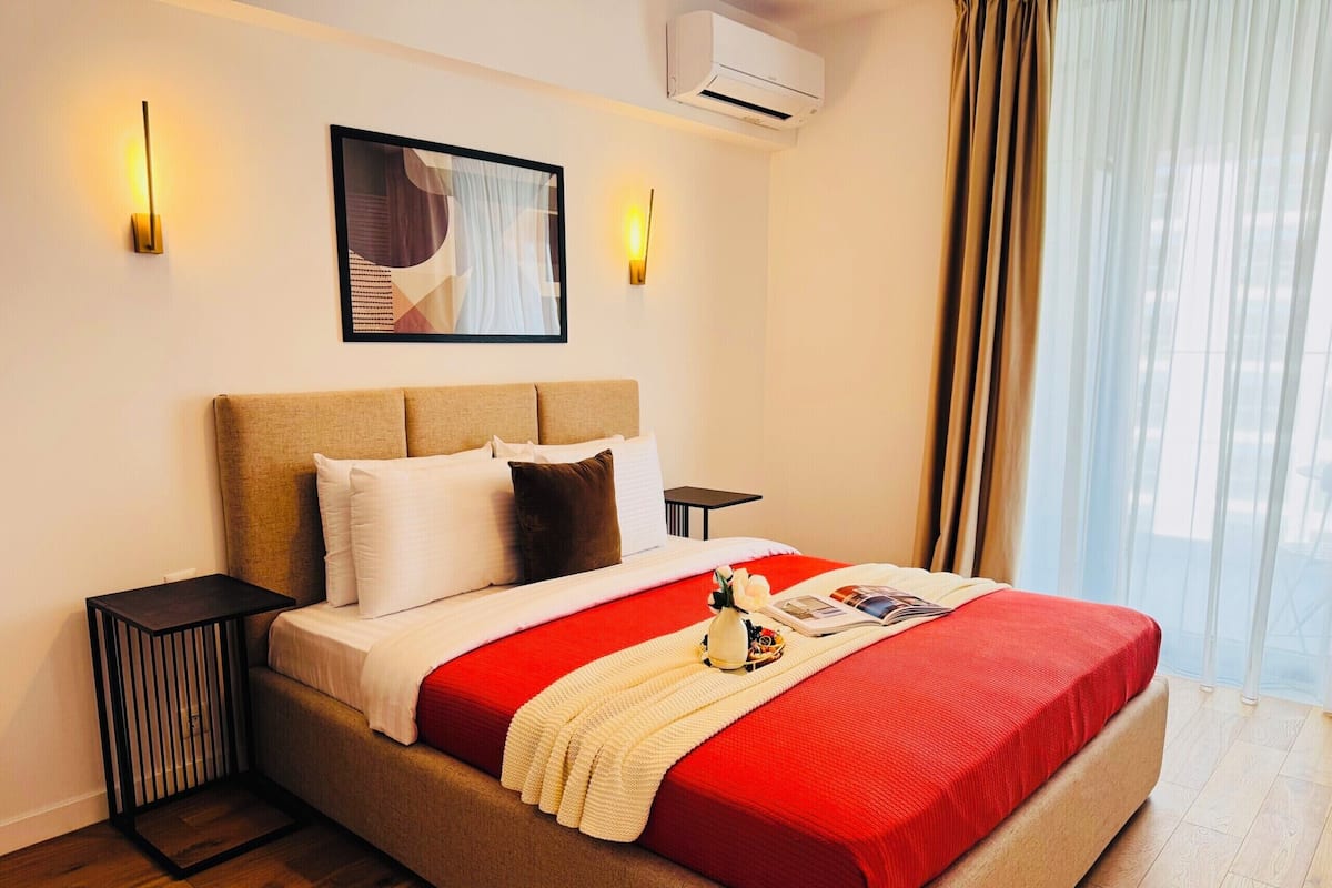 A hotel room with a bed, bedside tables, an air conditioning unit, a window with curtains, and a wall-mounted artwork.